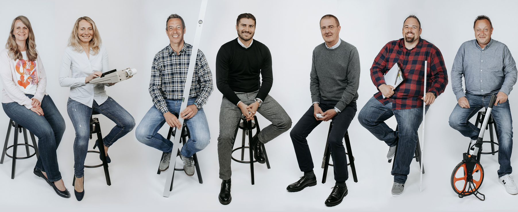 Das Büro Team der Firma Sechs Personen sitzen auf Hockern, lächelnd, in lockerer Kleidung vor neutralem Hintergrund. Es ist das Büro Team der Firma