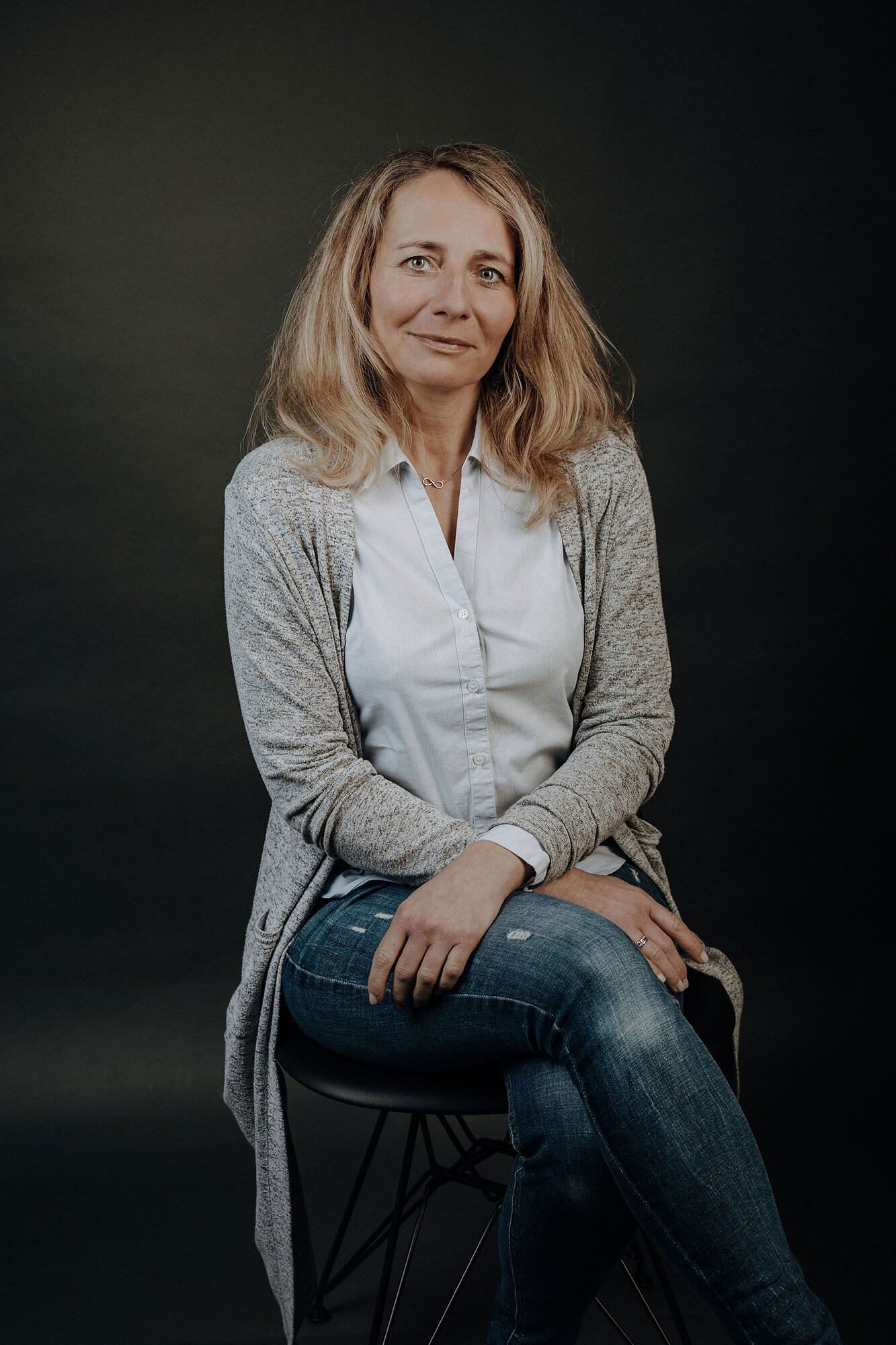 Teamassistentin Kerstin Nastke Frau mit blonden Haaren sitzt auf einem Stuhl, lächelt, trägt eine Bluse und Jeans.