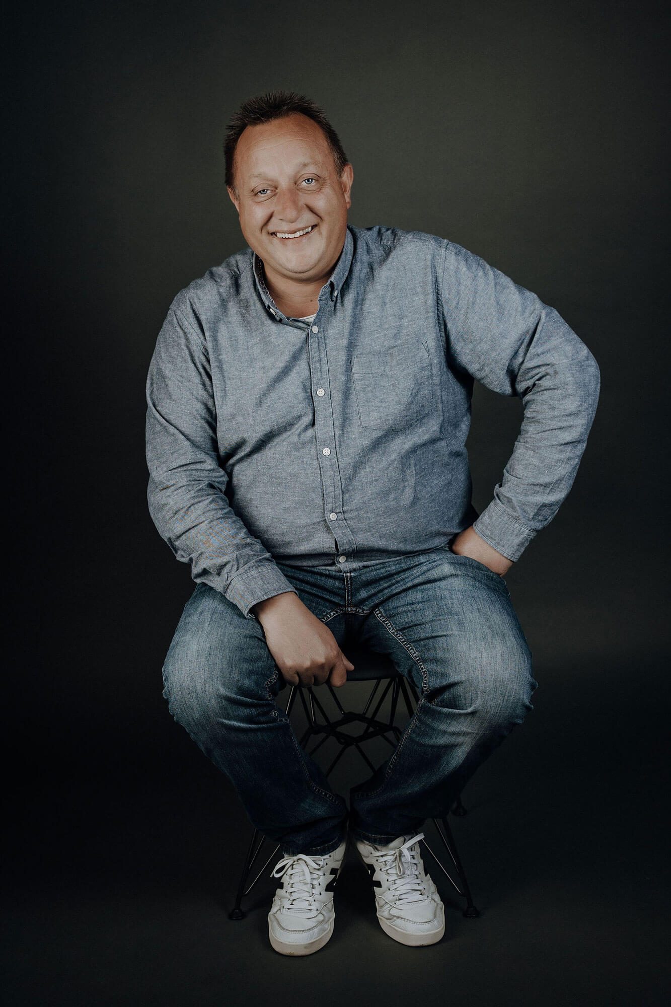 Bauleiter Tiefbau David Dohle Lächelnder Mann in einem blauen Hemd und Jeans, mit einer Hand auf dem Knie.