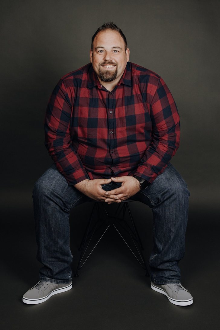 Bauleiter Tiefbau David Dohle Lächelnder Mann in einem blauen Hemd und Jeans, mit einer Hand auf dem Knie.