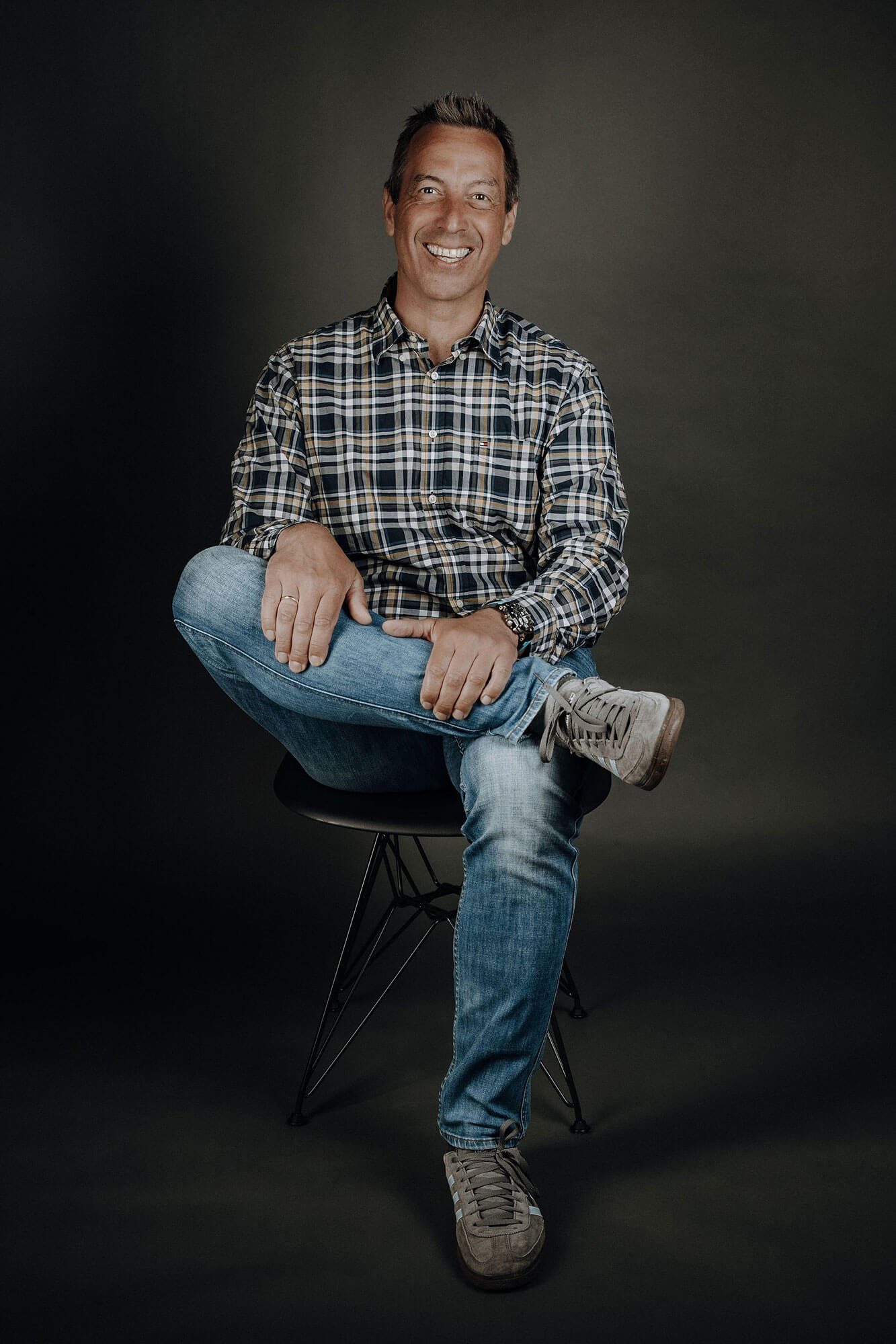 Bauleiter Hochbau Arndt Rousek Mann sitzt lächelnd auf einem Stuhl, trägt ein kariertes Hemd und Jeans.