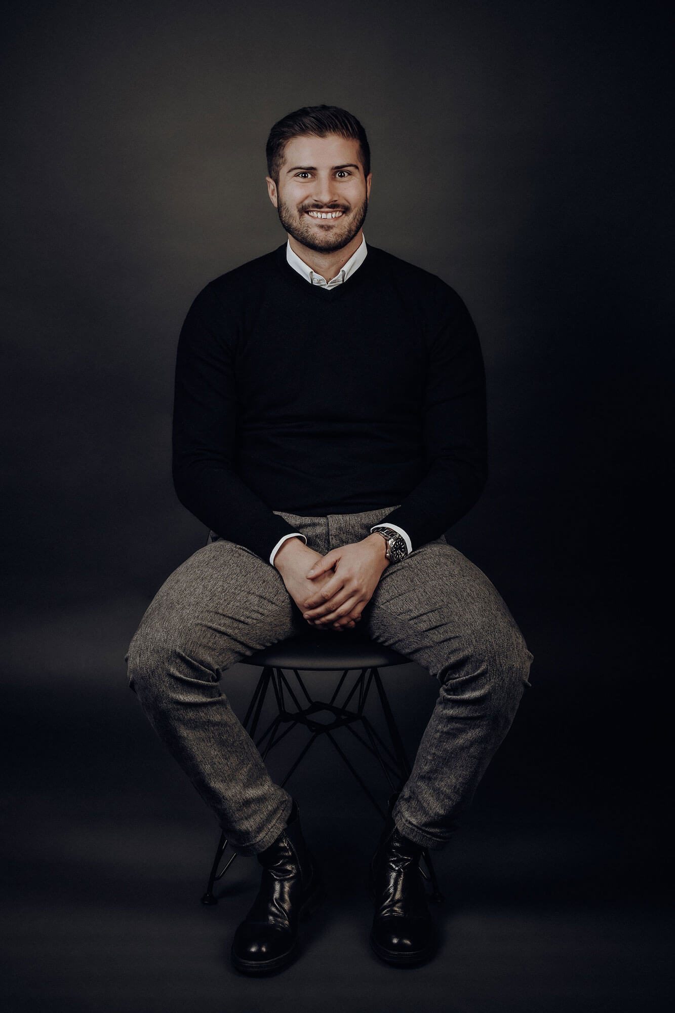 Geschäftsführer Niels Baldauf Geschäftsführer Niels Baldauf sitzt auf einem Hocker in eleganter Kleidung vor dunklem Hintergrund.