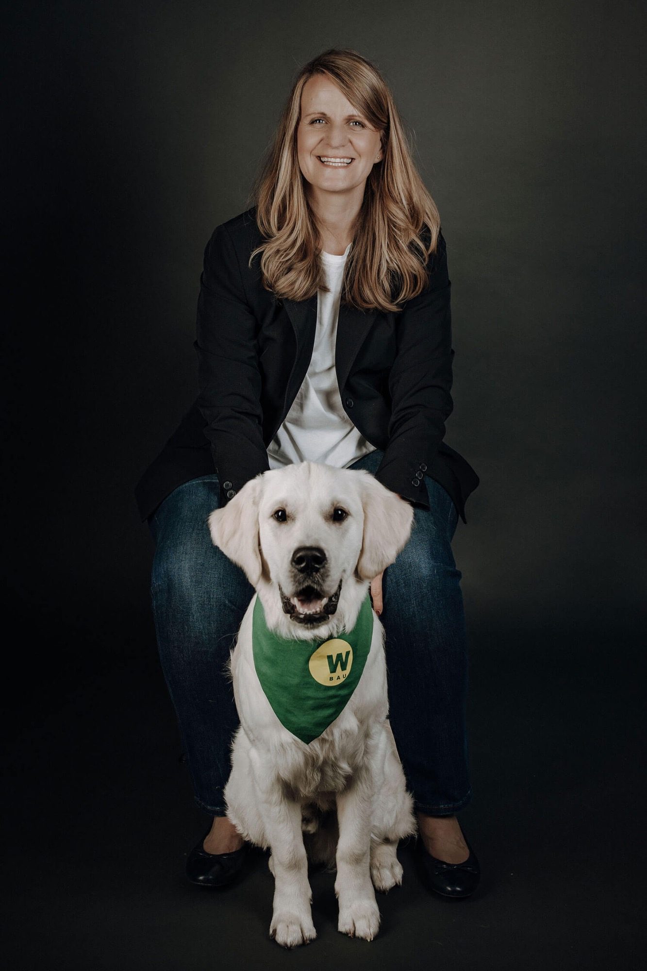 Buchhalterin Karola Sommerfeld Eine lächelnde Frau sitzt auf einem Stuhl . Vor ihr ein Golden Retriever mit grünen Halstuch.